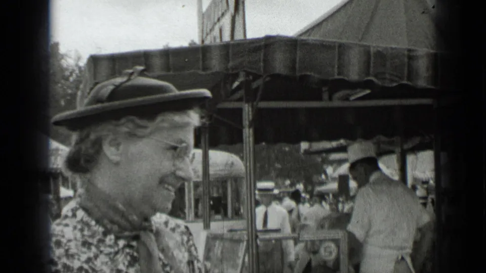 1939 New York home-movie frame of a woman in a hat and glasses smiling near a fair canopy and crowd.