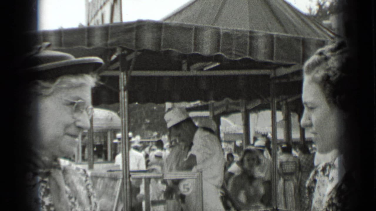 Two women stand near a covered fair stall with crowds behind them.