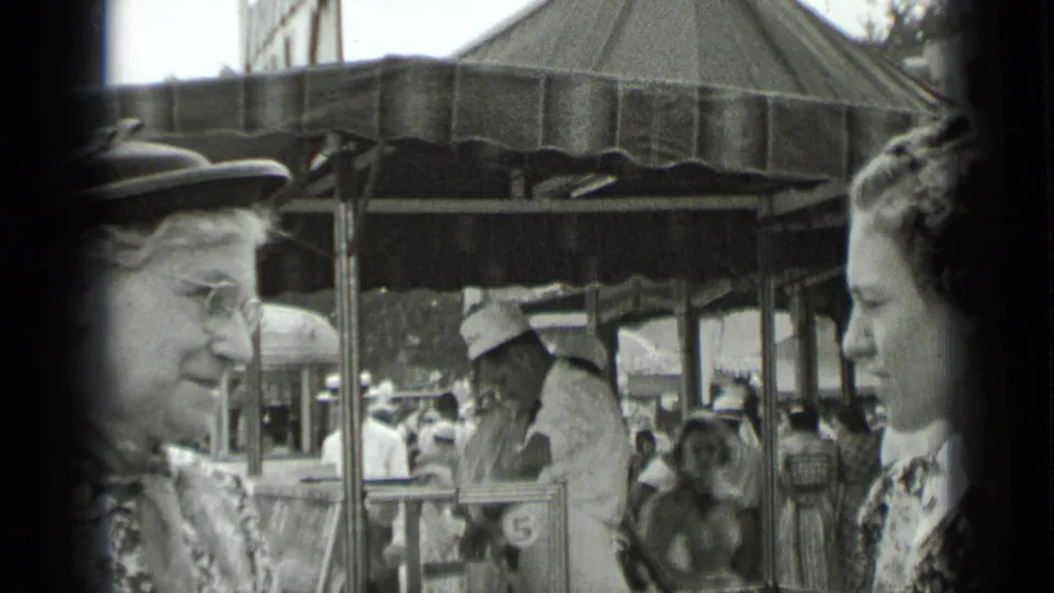 Two women in period dress stand near a covered fair stall in 1939 New York home-movie footage, with other fairgoers behind them.