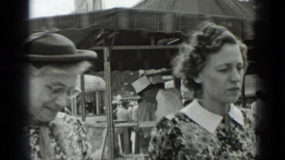 Two women in patterned dresses stand in the foreground of black-and-white 8mm footage, with fair visitors and a covered stall behind them.