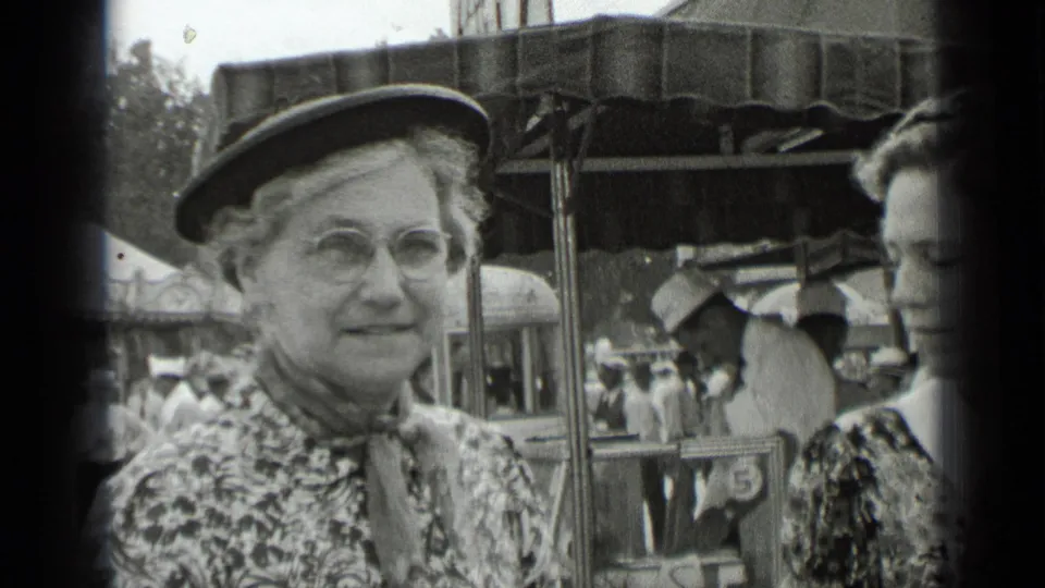 A woman wearing a dark hat and eyeglasses smiles near the camera, with a covered fair stall and visitors visible behind her.