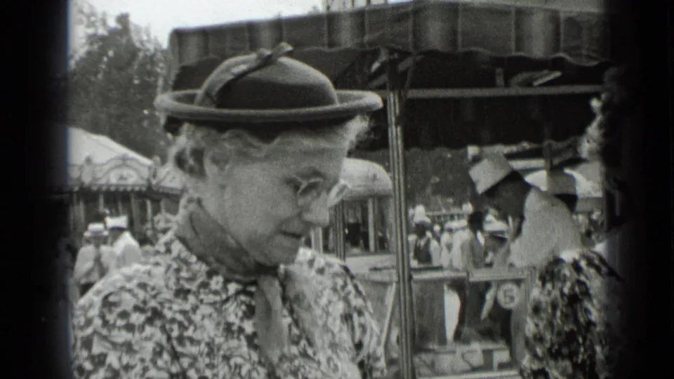 At a 1939 New York fair, an older woman in a patterned dress and brimmed hat is seen in profile near ride or stall structures and other visitors.