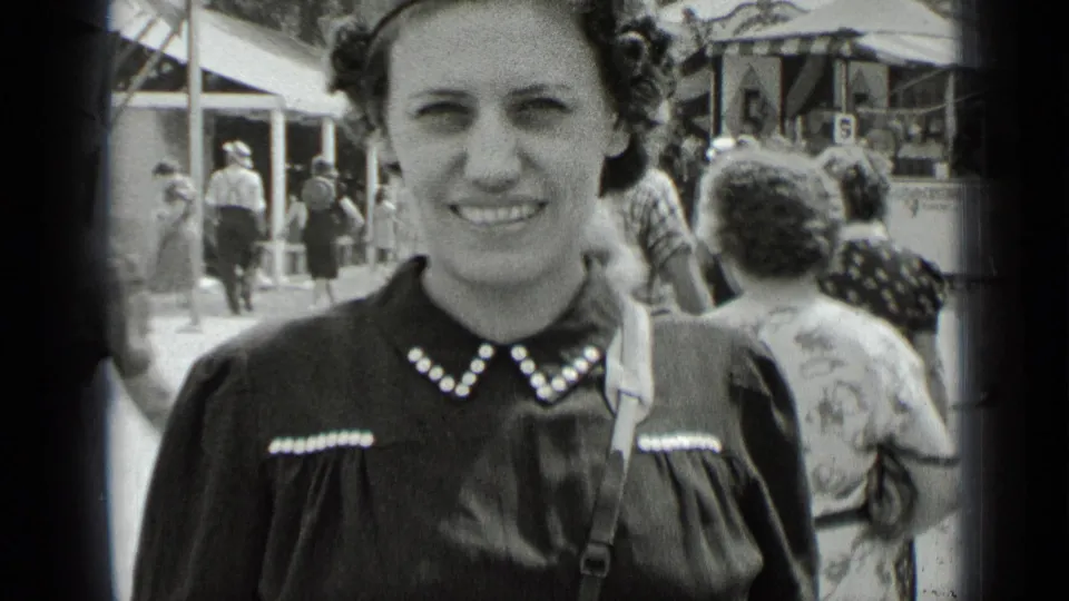 A young woman in a dark dress with a pearl-trimmed collar smiles at the camera amid a crowded 1939 New York fairground.