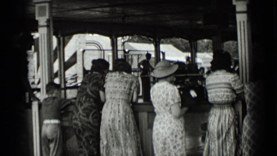 Several women in patterned dresses and hats stand with their backs to the camera at a shaded fair stall or ride counter in New York, 1939.