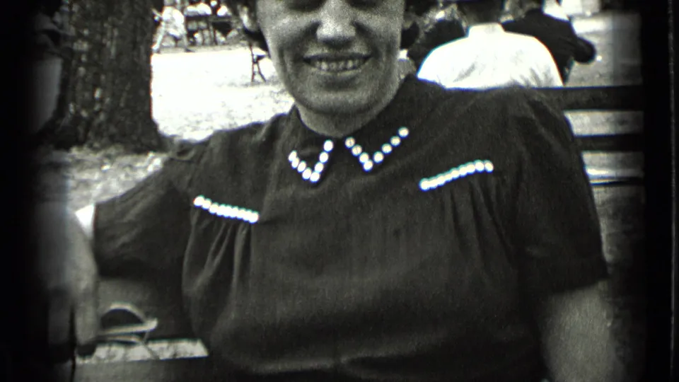 In 1939 New York home-movie footage, a woman smiles toward the camera while seated on a park bench in a dark dress with bright beaded trim.