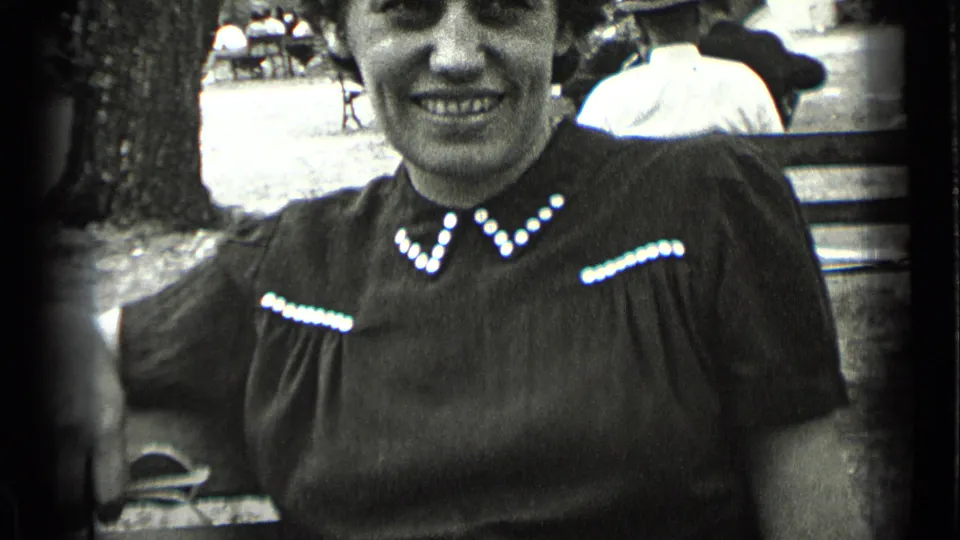 The woman remains seated on a New York park bench, smiling in soft black-and-white 8mm footage with trees and people behind her.