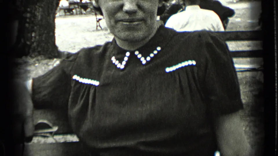 A close handheld 8mm frame shows the seated woman in a dark short-sleeved dress with white beaded trim, with a park bench and visitors behind her.
