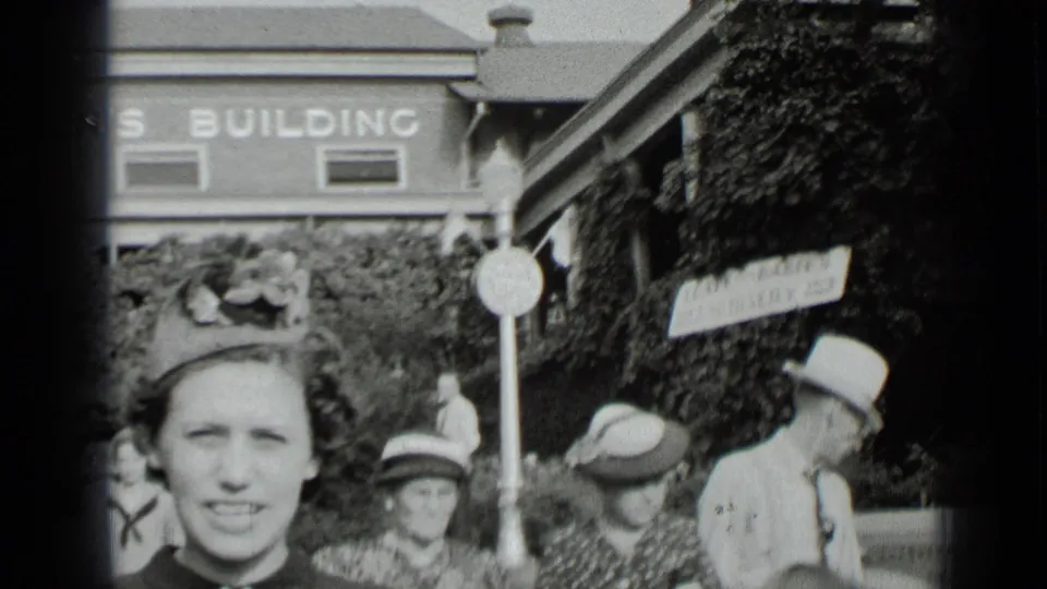 1939 New York home-movie frame showing adults in hats gathered near a building with a partial sign and ivy-covered wall.