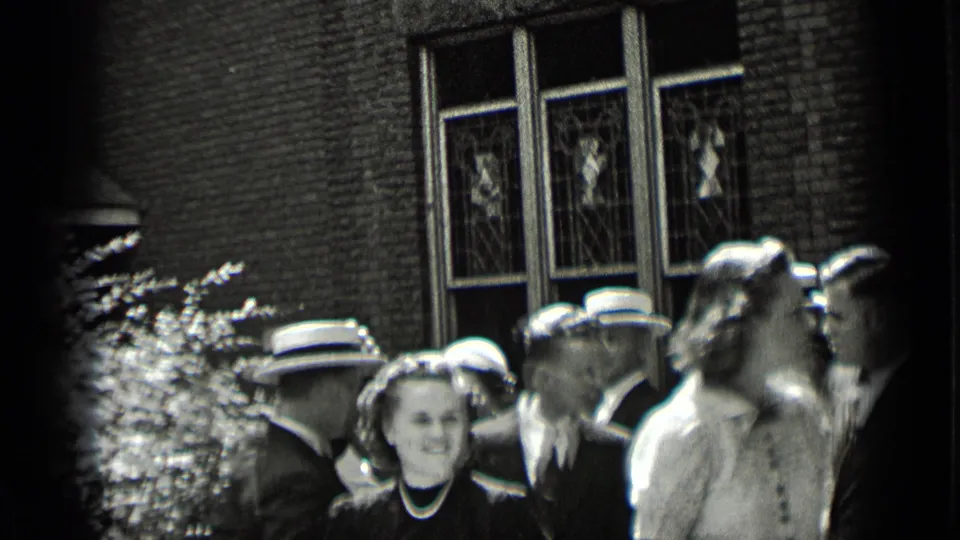 1939 New York home-movie frame of dressed adults in hats gathered outside a brick building with tall decorative windows.
