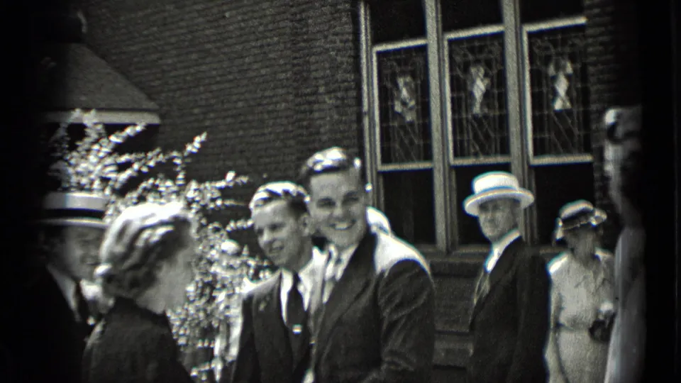 People in 1930s formal dress smile and converse outside a New York brick building with decorative windows.