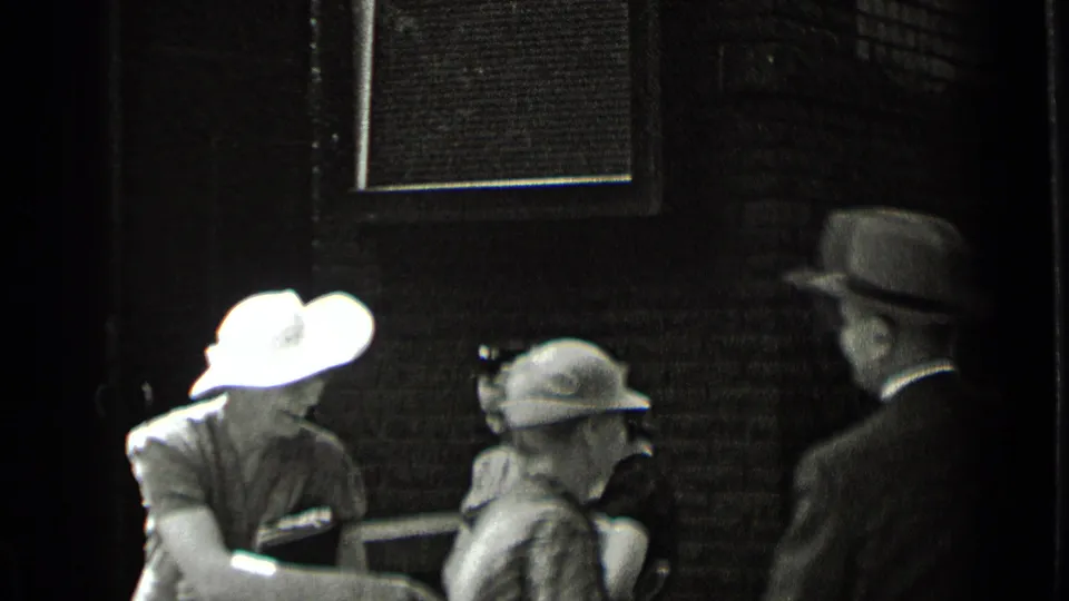 Two women in hats and dresses appear near a suited man outside the brick building in 1939 New York footage.