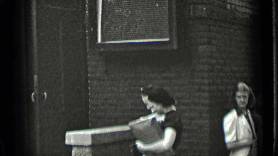 Outside the New York brick building, a woman carries a rectangular box or case as another woman stands nearby.