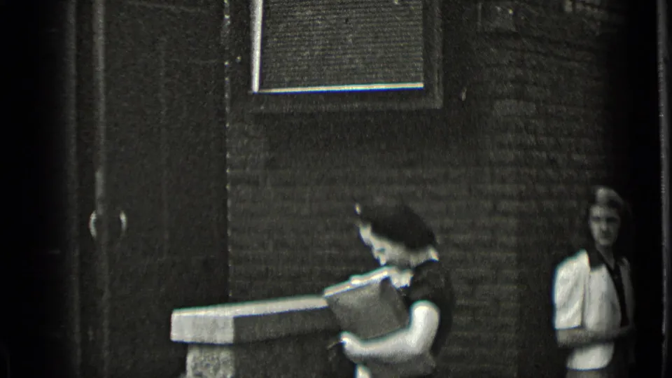 In 1939 New York home-movie footage, two women stand by a residential building entrance, with one holding a rectangular case or parcel.