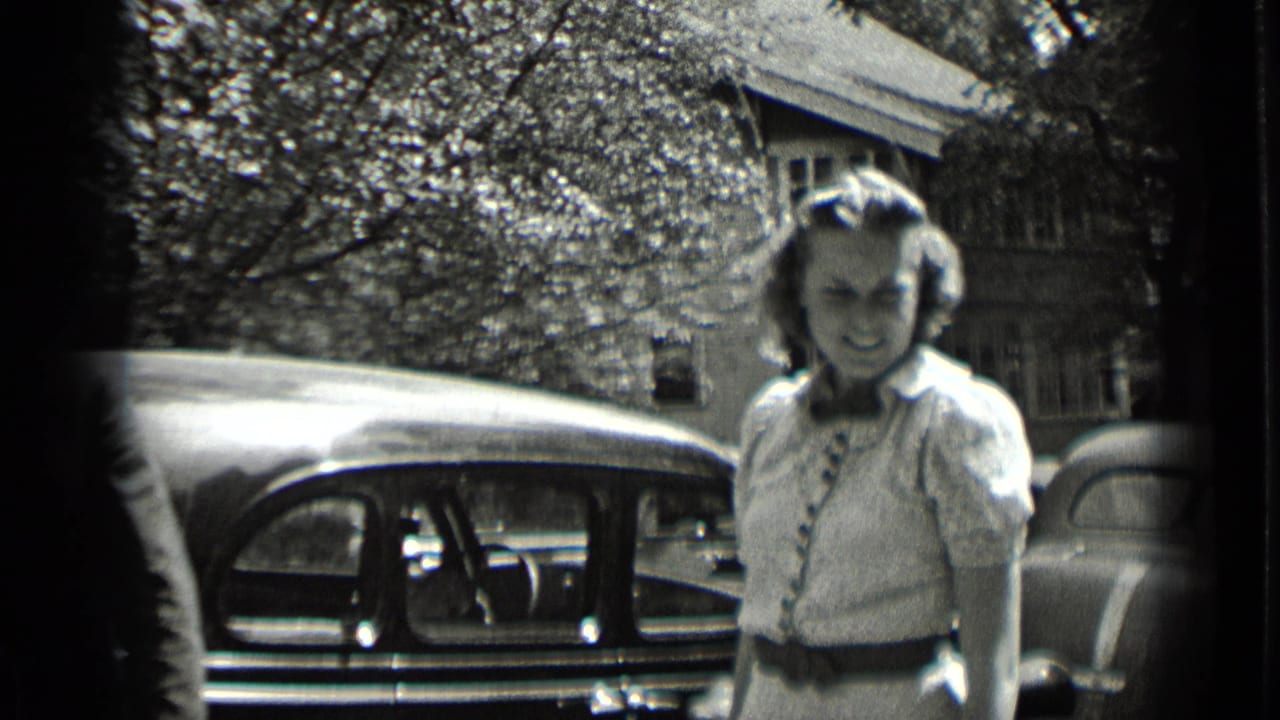 A young woman in a light belted dress stands on a residential street beside a parked rounded automobile in 1939 New York footage.