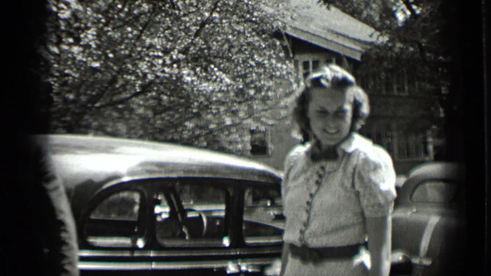 A young woman in a light belted dress stands on a residential street beside a parked rounded automobile in 1939 New York footage.