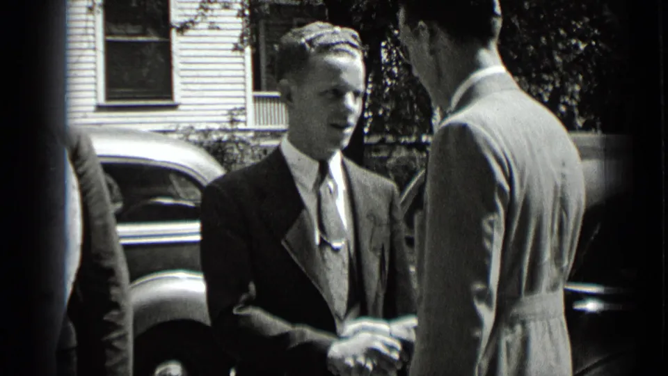 Two suited men shake hands on a New York residential street in 1939, with a parked car and house visible behind them.