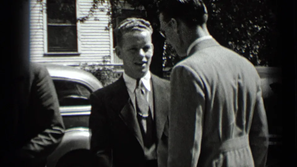 Two suited men stand close together on a residential street, with a parked car and house behind them in 1939 New York home-movie footage.