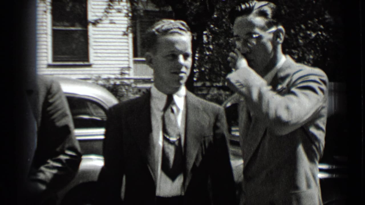 Two men in suits stand outdoors near a parked car and house siding.