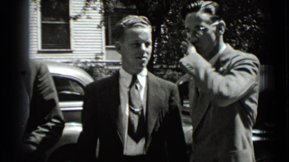 In 1939 New York home-movie footage, suited men stand in sunlight near a parked vintage car and residential-looking siding.