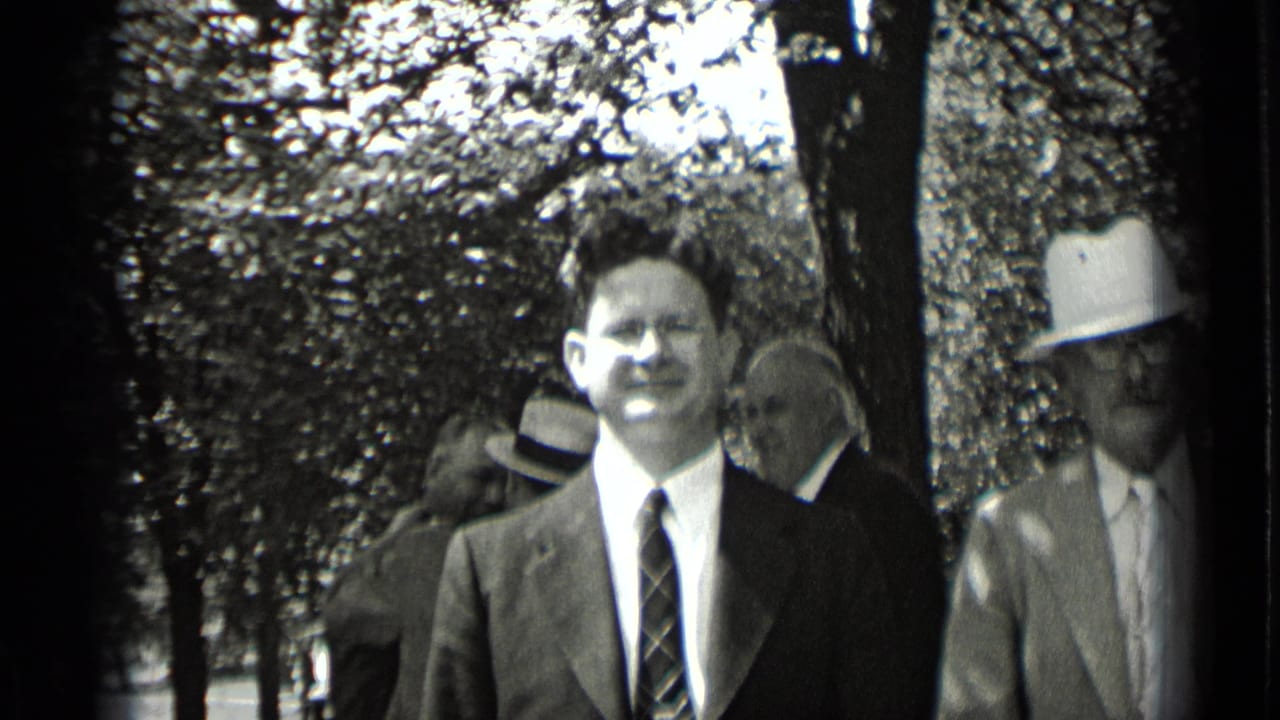 A man in glasses faces the camera while other suited men stand behind him.
