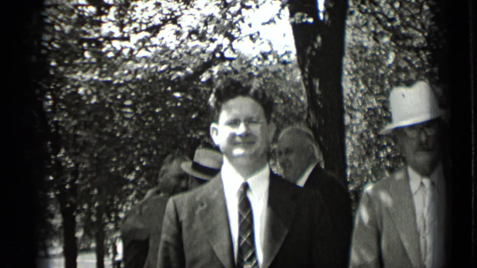 A suited man wearing glasses looks toward the camera in a leafy New York outdoor setting, with other men and hats behind him.