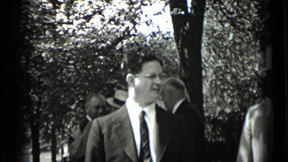 The man in glasses appears mid-conversation or mid-expression while suited men and hat-wearing figures gather under trees.