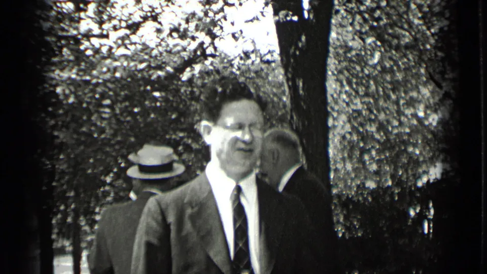 In the closing frame, a suited man with glasses stands foregrounded beneath trees while other men remain in conversation behind him.