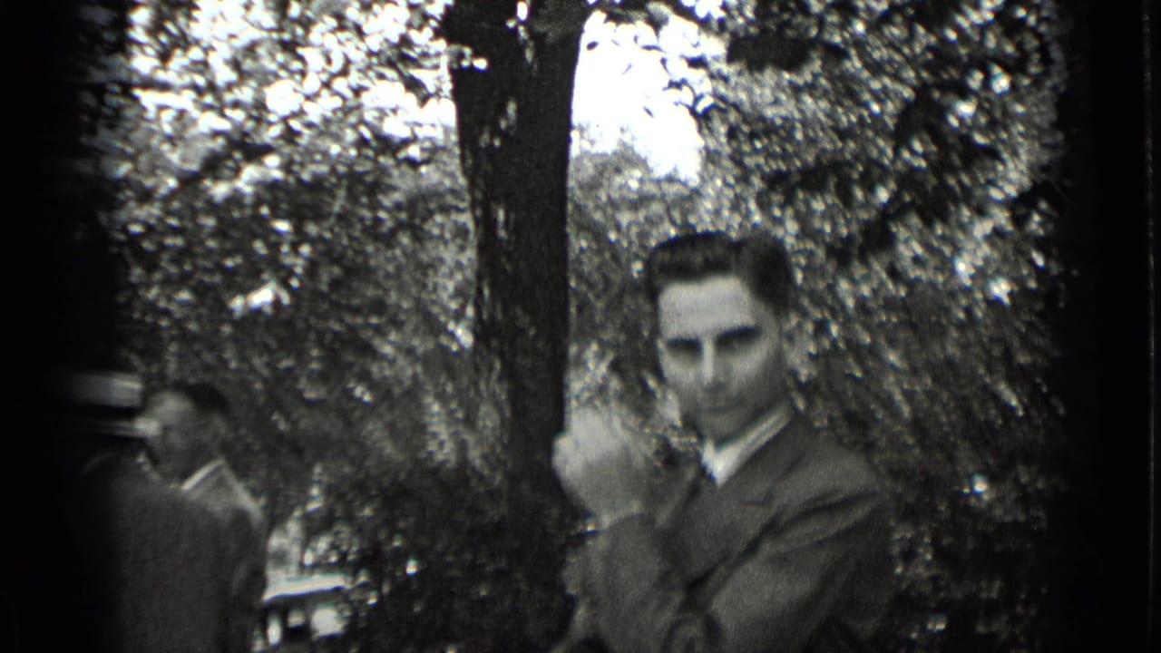 Man in a suit looks toward the camera beside trees, with other adults partly visible.