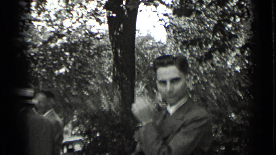 In 1939 New York home-movie footage, a suited man faces the camera while other adults stand near trees in soft focus.
