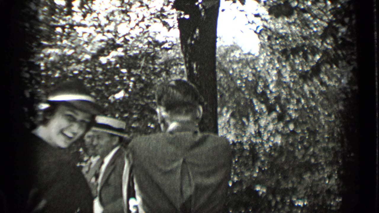 A woman in period dress and a brimmed hat smiles toward the lens as men in suits stand nearby among trees.