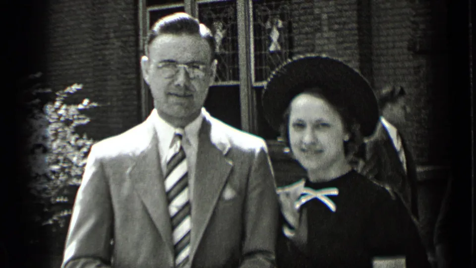 A man in a light suit and striped tie stands with a woman in a dark wide-brimmed hat before a brick building in New York footage.