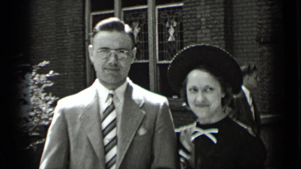 In 1939 New York home-movie footage, a suited man and a woman in a dark dress and wide-brimmed hat face the camera outside brick walls and windows.