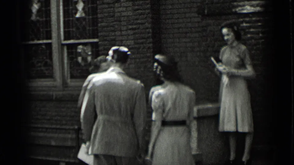 Adults in period clothing stand and turn near stained-glass windows on a brick exterior, with one woman holding a small book or folded item.