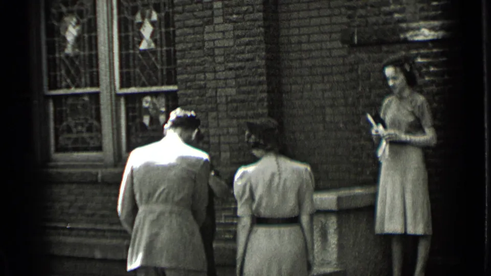 A man in a suit and a woman in a belted dress move away from the camera as another woman stands beside the brick wall holding an item.