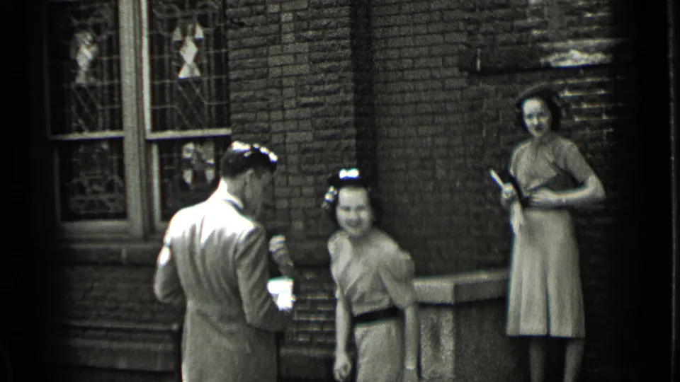 A suited man and a woman in a light dress appear to laugh or talk near the brick building while another woman stands to the side holding an item.
