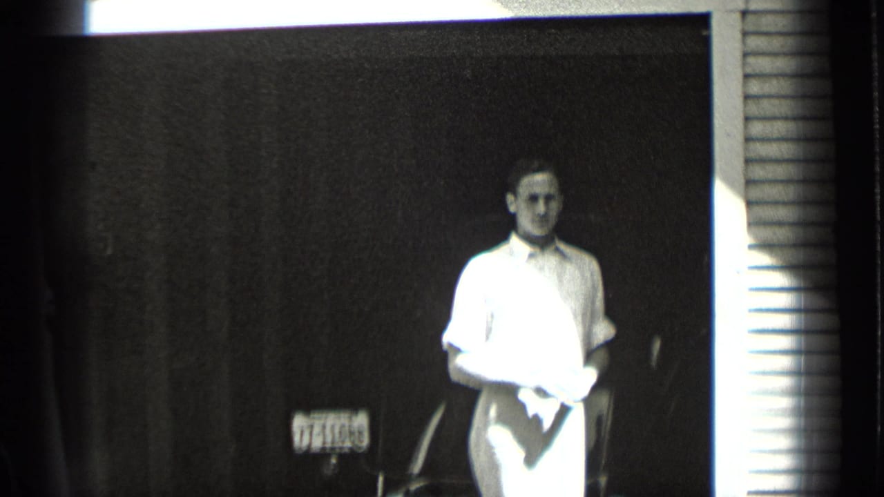 Young man in a white shirt stands in a dark doorway holding a cloth near a visible license plate.