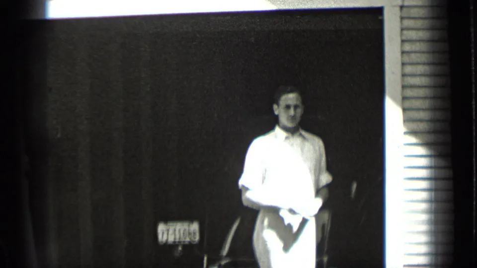 In supplied 1939 New York home-movie footage, a young man stands at a doorway holding a cloth, with siding and a license plate behind him.