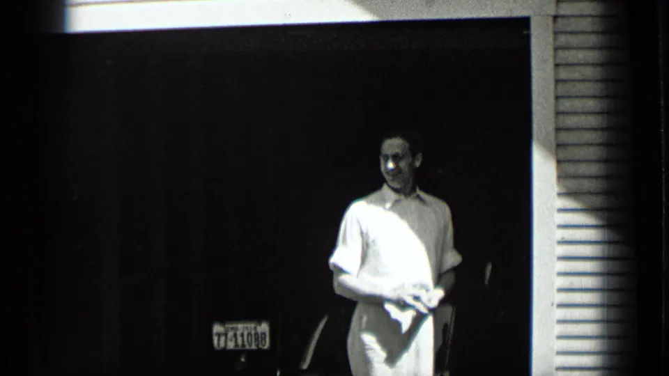 A smiling young man in light clothing stands near a dark doorway in the supplied New York home-movie scene, with a plate visible low in frame.