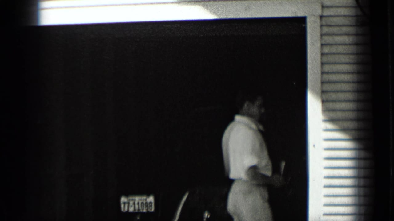 Man standing in a garage doorway beside part of a car and a visible license plate area.