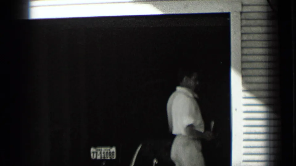 1939 New York home-movie frame showing a man in a garage doorway, partly turned near a car, with siding and a license plate area visible.