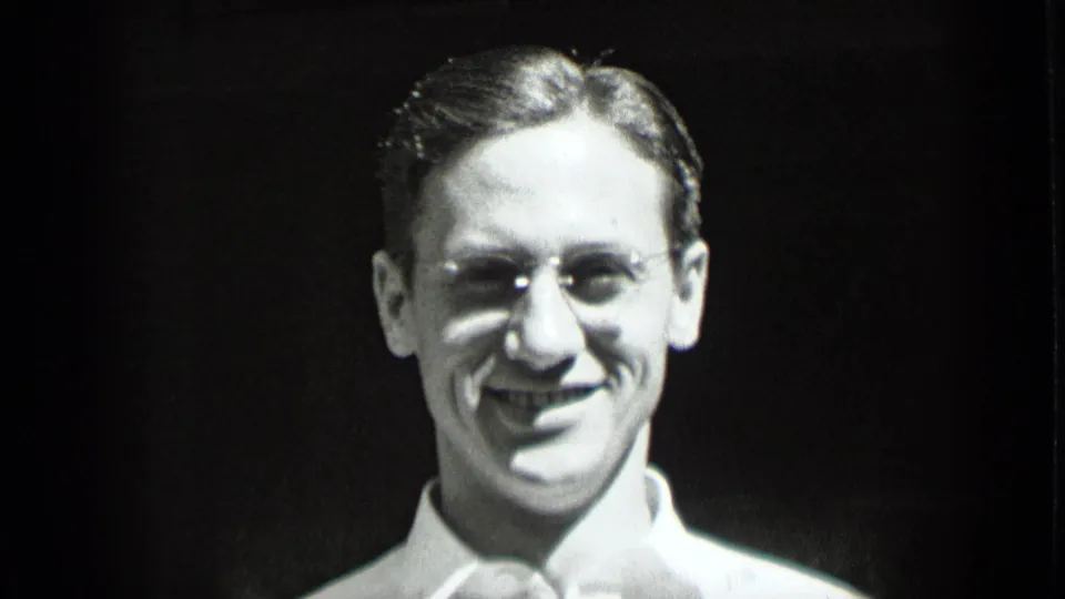 1939 New York black-and-white close-up of a smiling young man wearing round glasses and a white collared shirt against a dark background.