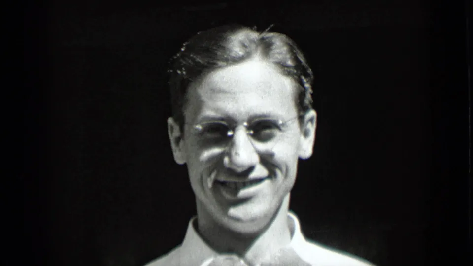 1939 New York black-and-white frame of the smiling man centered in close-up, wearing glasses and a white collared shirt.