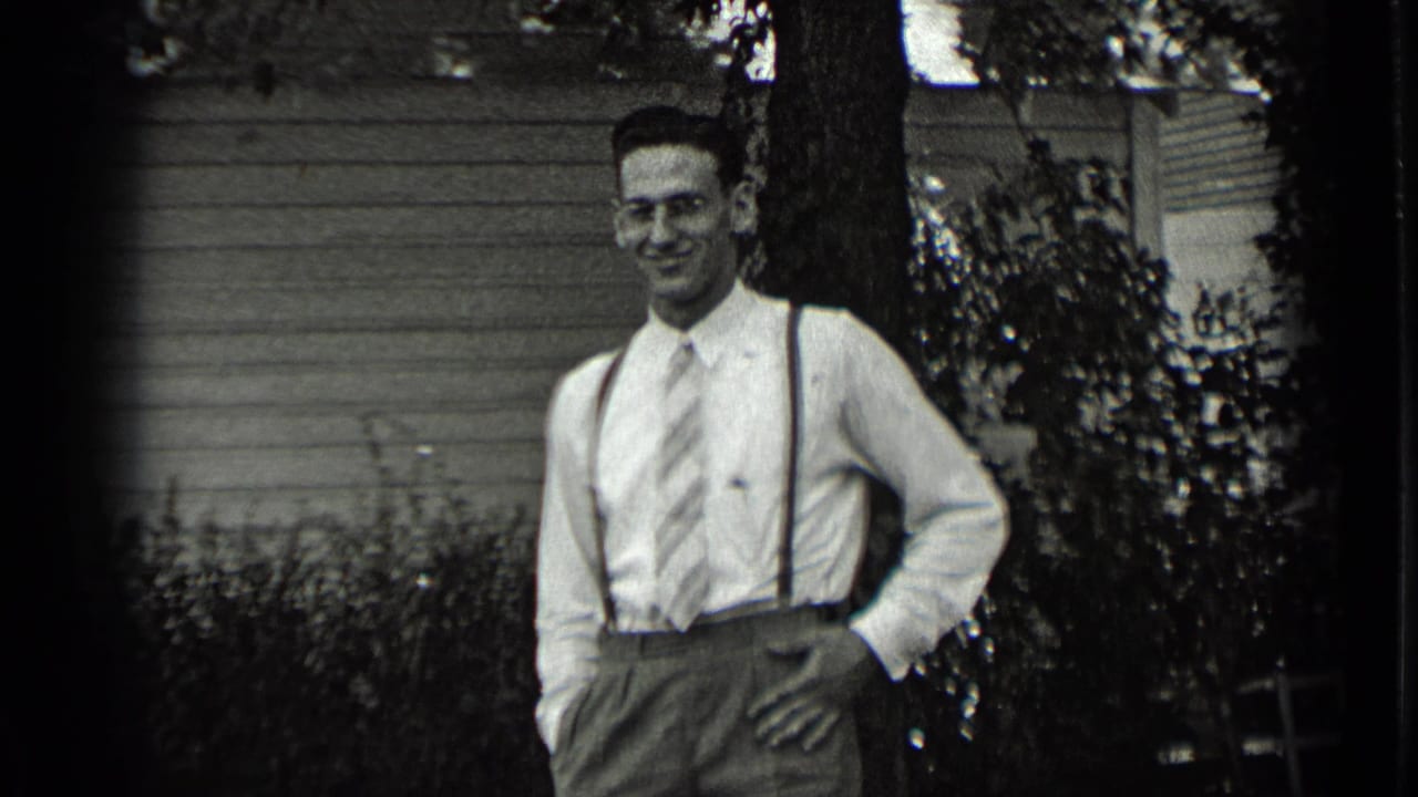A young man in 1939 New York stands in a garden setting wearing a dress shirt, striped tie, suspenders, and high-waisted trousers.