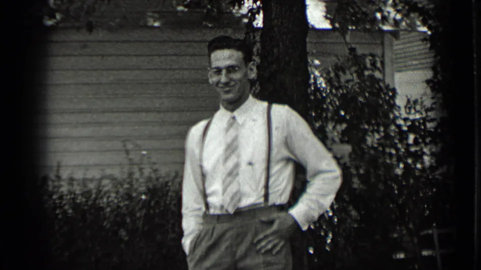 A young man in 1939 New York stands in a garden setting wearing a dress shirt, striped tie, suspenders, and high-waisted trousers.