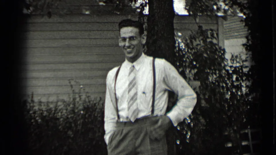 The man smiles toward the camera in a 1939 New York garden scene, with suspenders, striped tie, shrubs, and house siding visible.
