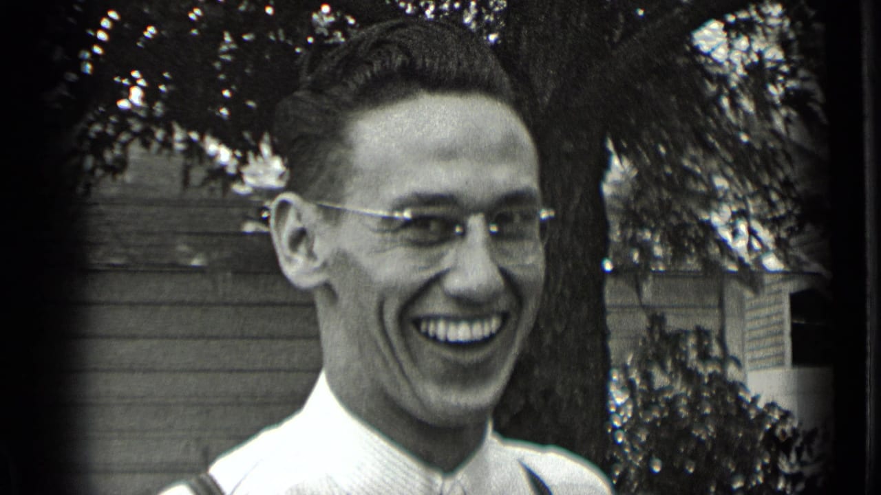 Smiling young man with glasses standing outdoors near trees and a fence.