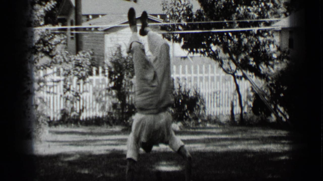 The boy remains inverted on the lawn, framed by a white fence, trees, shrubs, and a house exterior in New York home-movie footage.