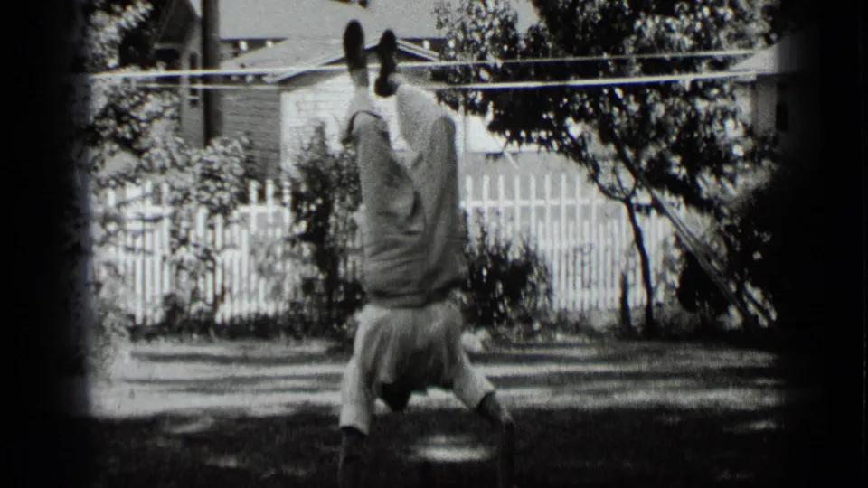 The boy remains inverted on the lawn, framed by a white fence, trees, shrubs, and a house exterior in New York home-movie footage.