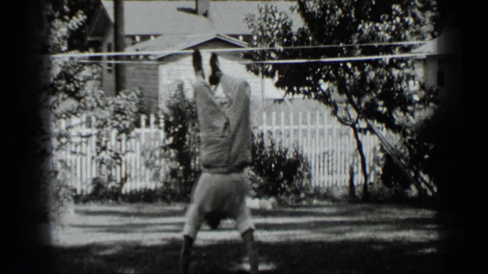 A boy in light clothing continues practicing a handstand on a backyard lawn, with residential fencing and plants behind him.
