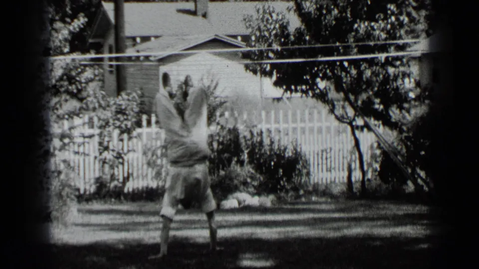 The boy appears to come down from the handstand on the lawn, with trees, a white fence, shrubs, and house exterior visible.